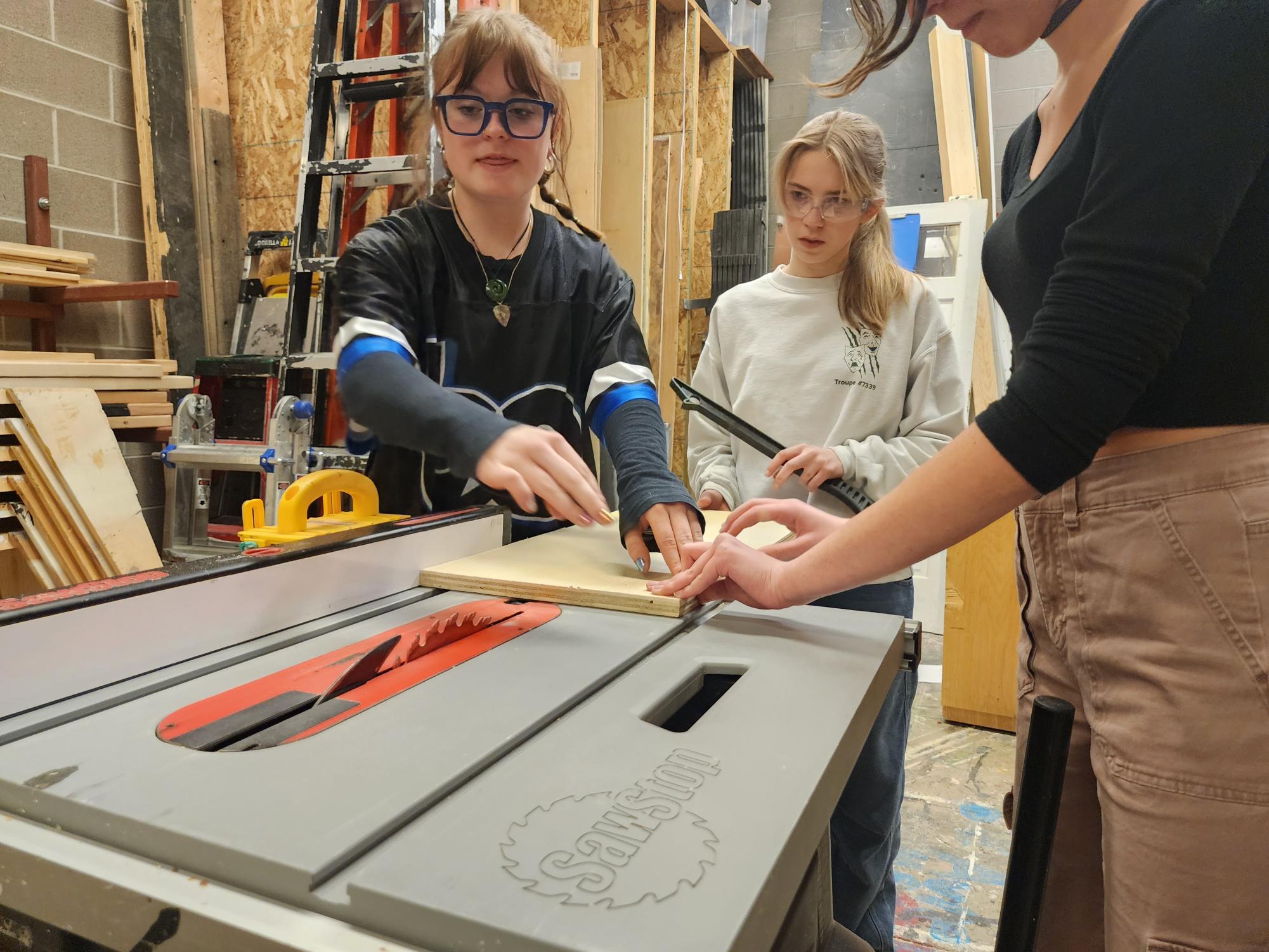 Brynn Olsen assists Chloe Cropp and Stella Powers with how to push wood on the table saw.